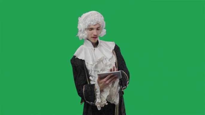 穿着黑色复古西装和假发的朝臣绅士肖像，用平板电脑戴着耳机欣赏音乐。年轻人在工作室摆姿势，绿屏背景。特
