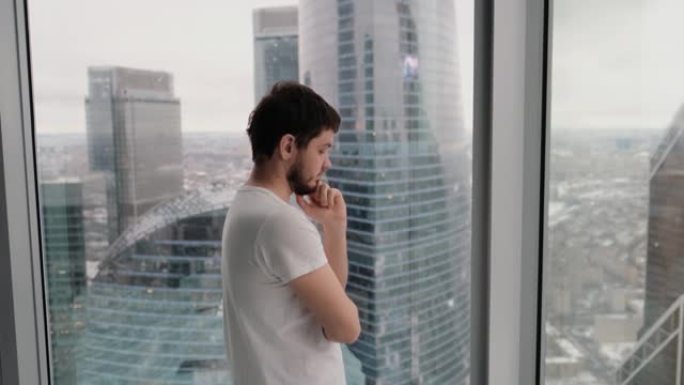 年轻人在顶层俯瞰城市的现代公寓里思考并看着窗外