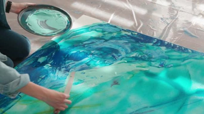 女性艺术家使用艺术调色板和抹刀在画布上应用蓝色的特写镜头。熟练的年轻女性在工作场所创造抽象模式。