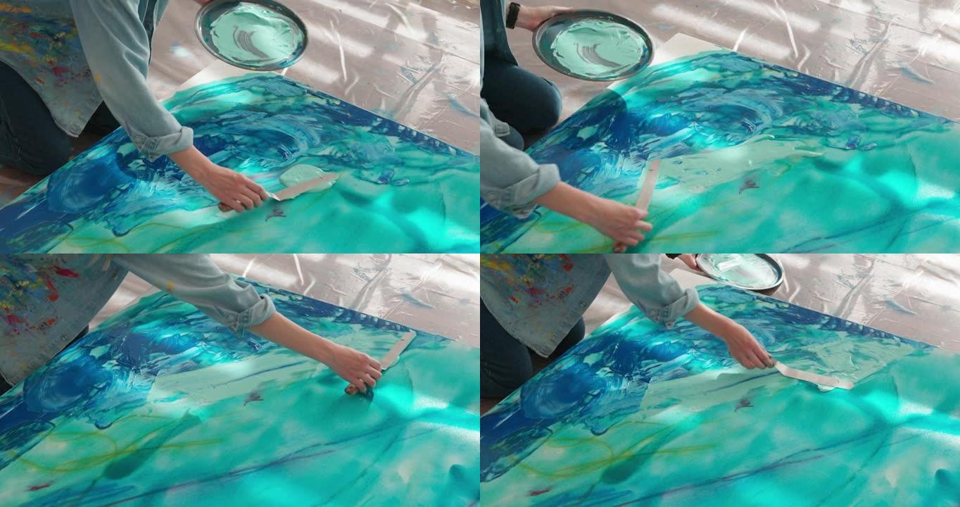 女性艺术家使用艺术调色板和抹刀在画布上应用蓝色的特写镜头。熟练的年轻女性在工作场所创造抽象模式。