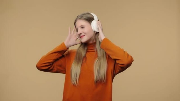 大型无线白色耳机中享受音乐的时尚模特肖像。穿着橙色毛衣的长发年轻时尚女孩在棕色工作室背景下摆姿势。特