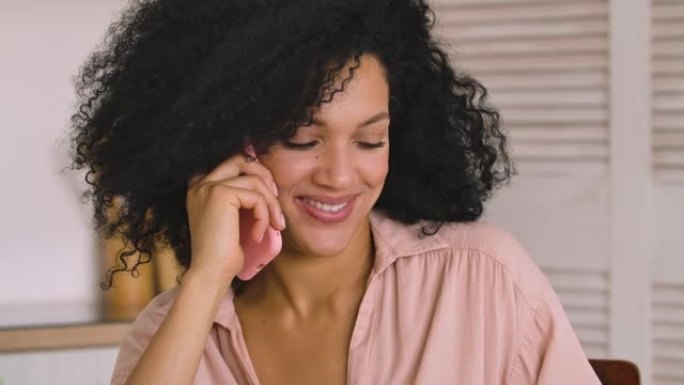可爱的非洲裔美国女人在手机上聊天，微笑着开心。黑色黑发女人，卷发，坐在明亮的房间里摆姿势。特写。慢动