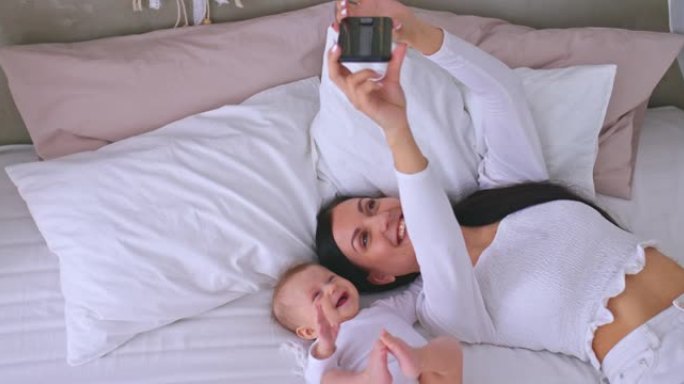 美丽的妈妈用手机和宝宝自拍，躺在床上微笑。年轻的母亲与她的小女儿在智能手机上合影。4k，普洛雷斯