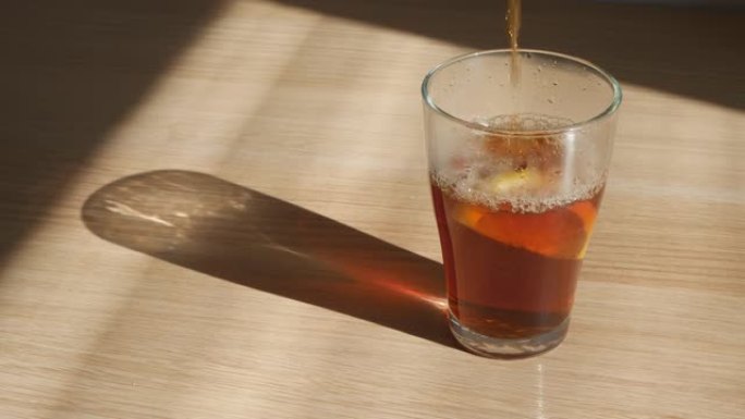 美丽的茶被倒入一个大玻璃杯中。玻璃杯的底部是一个柠檬，它在桌子上，阳光照在上面。茶的美丽色彩在阳光下