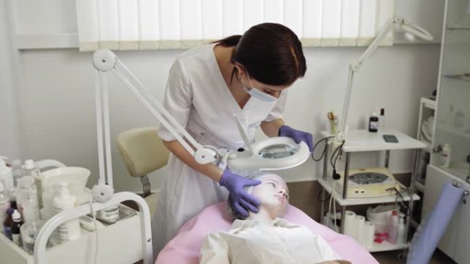 护肤概念。美容办公室的女医生检查年轻漂亮的少女戴着一次性帽子的面部皮肤，有痤疮问题的皮肤。皮肤护理程