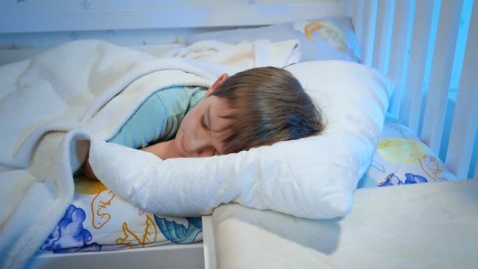 晚上穿着睡衣的小男孩在白色毯子下睡在床上的视频。孩子晚上休息和做好梦