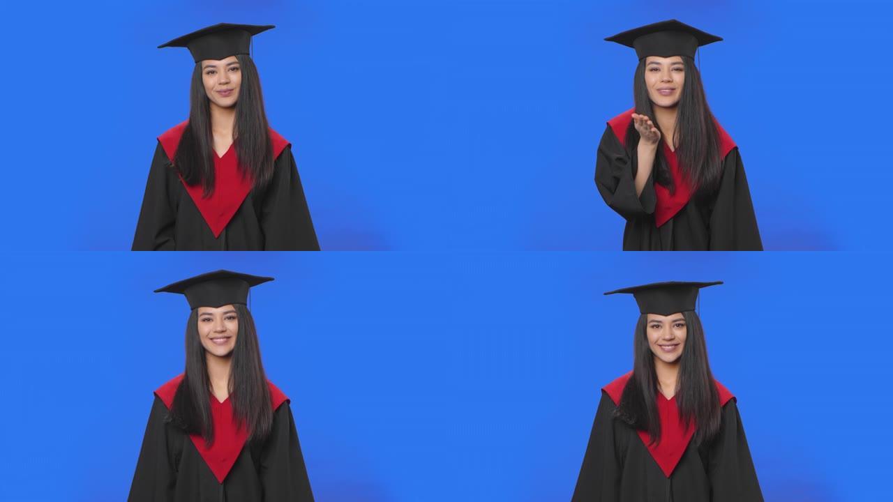 穿着帽子和长袍毕业服装的女学生的肖像微笑和吹吻。年轻的黑发女人在工作室摆姿势，蓝屏背景。特写。慢动作