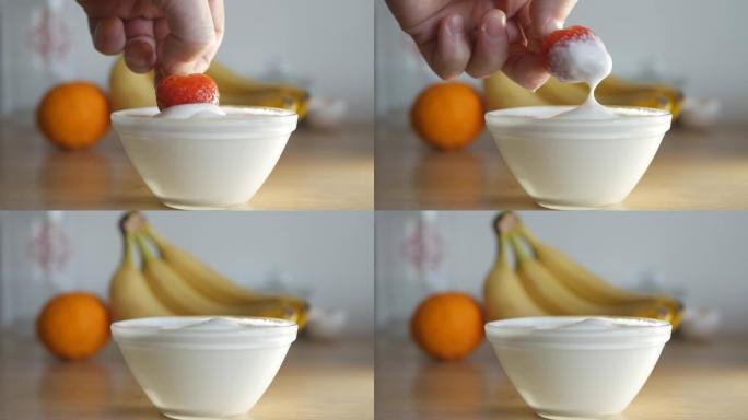 一个人的手将成熟的红色草莓浸入一碗鲜奶油中。背景是水果香蕉和柑橘。关闭4k