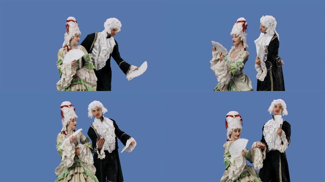 穿着历史悠久的老式服装和假发的朝臣女士和绅士的肖像挥舞着扇子。年轻的女人和男人在工作室摆姿势，蓝屏背
