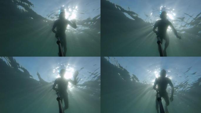 一个戴着面具和通气管的年轻运动男子在水下深处用自拍杆游泳。阳光透过水
