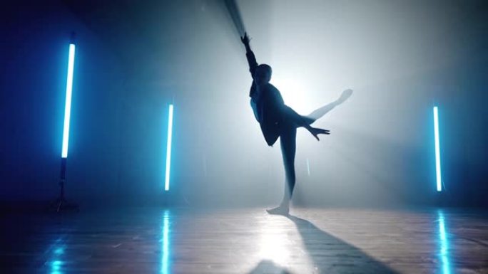 聚光灯背景下当代舞者的剪影。专业芭蕾舞演员在烟雾舞台上，配有蓝色灯，独奏表演。