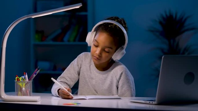 一个戴着耳机的女孩在听音频课时做笔记，看着笔记本电脑，点了点头。一个女孩在台灯的灯光下坐在桌子旁。特