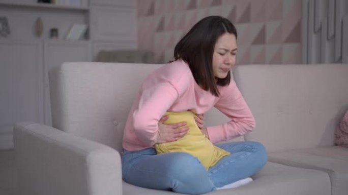 压力很大的亚洲女士坐在沙发上，抱着抱枕，肚子疼。