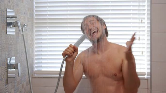 快乐的年轻人唱歌洗澡。微笑的大胡子肌肉男人在温水下洗衣服，听音乐，享受浴室里的卫生习惯。