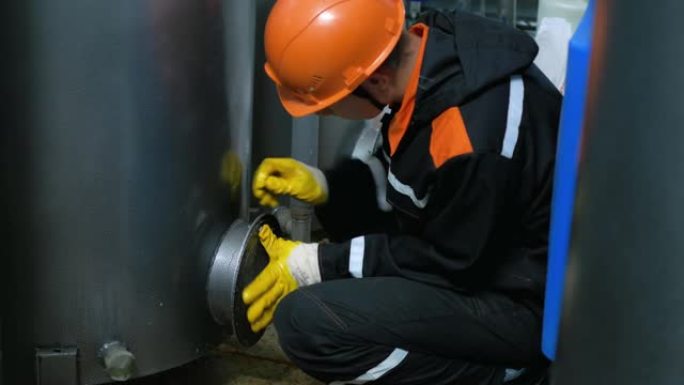 一名戴着头盔和手套的工人，拧下水箱舱口并更换吸附材料，以过滤垂直水箱中的水。工业专业。危险的工作