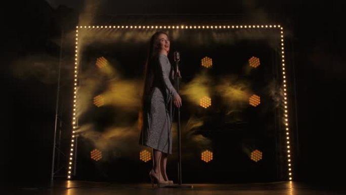 一位迷人的女歌手在灯光明亮的黑暗舞台上表演个人音乐会。穿着闪亮连衣裙的黑发女人触摸老式麦克风，并随着