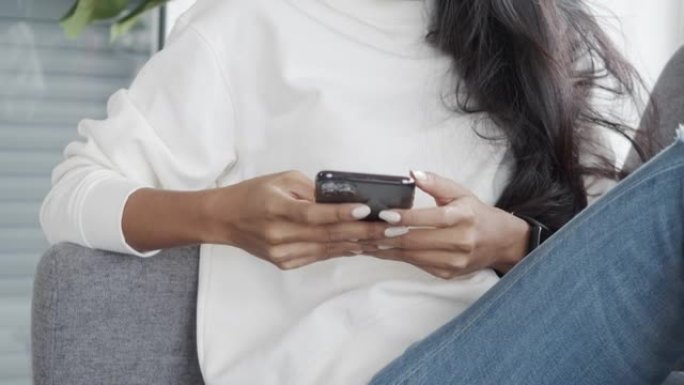 亚洲妇女坐在家里客厅的沙发上与朋友在线聊天。在智能手机上关闭女性打字信息。