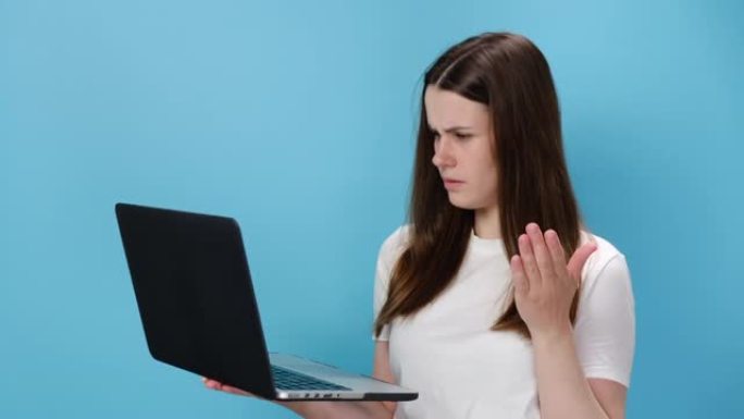 忧心忡忡的20岁年轻女孩在笔记本电脑上张开双手，问什么脏话显示拇指朝下，穿着白色t恤，孤立在蓝色工作