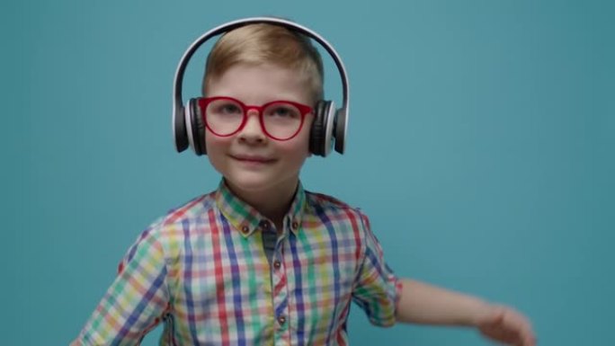 戴着眼镜和耳机的微笑可爱的男生在慢动作的蓝色背景上跳舞。快乐的孩子看着相机跳舞。