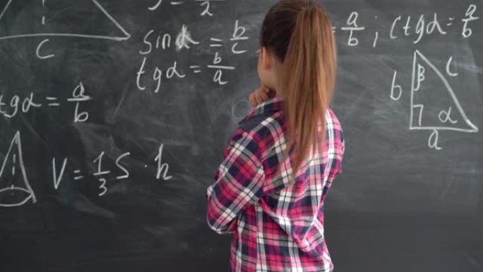穿着衬衫的高加索女孩女学生站在带有公式的粉笔板的背景下。试着解决这个等式，微笑着。复制空间。