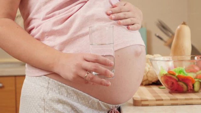 孕妇拿着一杯水，抚摸大肚子的特写镜头。怀孕期间健康生活方式、营养和水合的概念