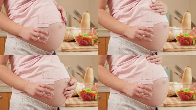 孕妇拿着一杯水，抚摸大肚子的特写镜头。怀孕期间健康生活方式、营养和水合的概念