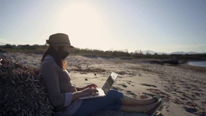 电影拍摄的年轻企业家在日落时在海水附近的大自然中工作的背光效果坐在沙滩上的沙滩上，使用笔记本电脑处理