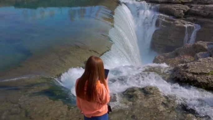 穿着蓝色牛仔裤和粉色运动衫的年轻红发女旅行者在阳光明媚的日子里用智能手机拍摄瀑布和峡谷