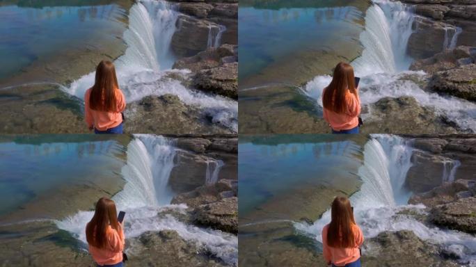 穿着蓝色牛仔裤和粉色运动衫的年轻红发女旅行者在阳光明媚的日子里用智能手机拍摄瀑布和峡谷
