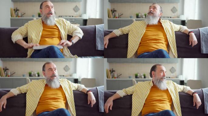 倾斜中等镜头的高级男子灰胡子完成工作坐在家里沙发上的笔记本电脑。男人休息，关闭笔记本电脑，叹息并向后