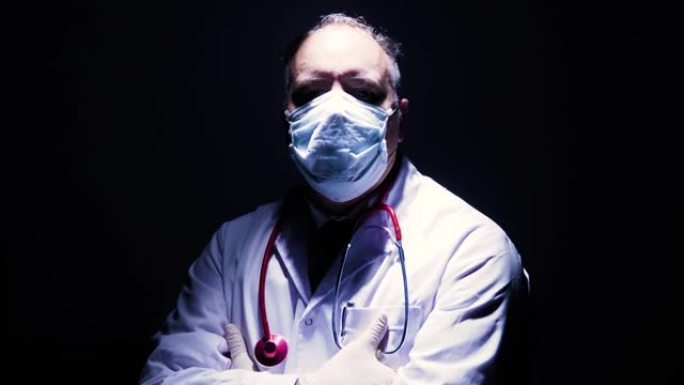 戴着口罩和手套的医生肖像在黑暗中看着相机，双手交叉。他是一位英雄，就像护士医生病毒学家一样，与冠状病