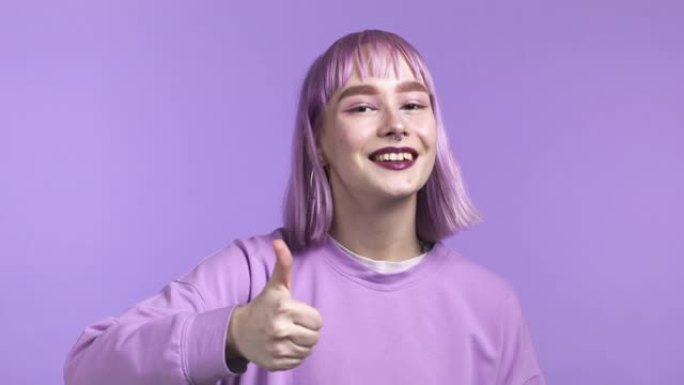 漂亮的女人露出拇指，就像在紫罗兰工作室背景上签名一样。染成紫色头发的积极年轻女孩对镜头微笑。赢家。成
