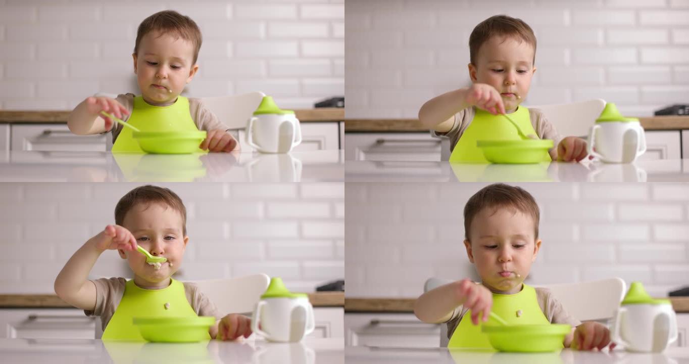 坐在桌子旁，用勺子吃燕麦片的可爱的小男孩的变焦镜头。宝宝在家心甘情愿地吃东西。概念快乐童年