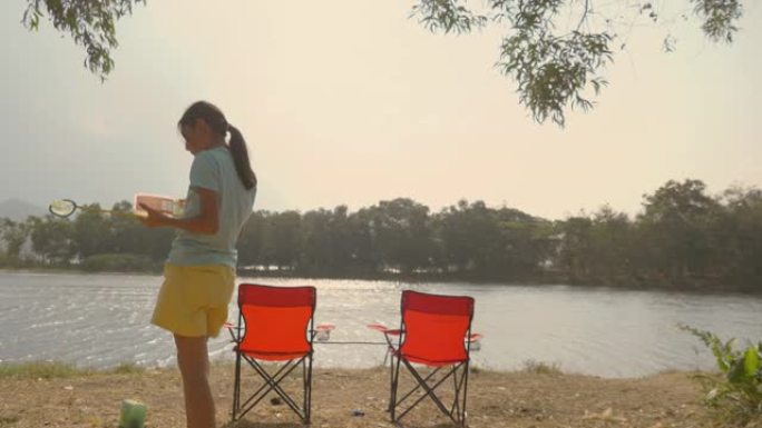 亚洲女孩在湖边放松户外附近的红色野营椅与太阳灯，生活方式的概念。