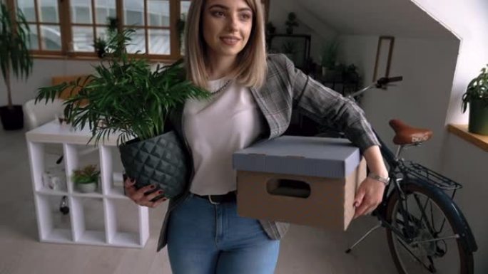 快乐的年轻女子带着植物和盒子搬进了新的家庭办公室设置