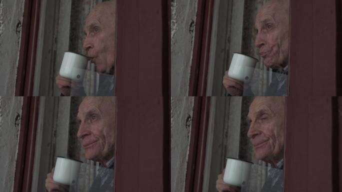 悲伤的老人站在窗玻璃后面，从杯子里喝茶。