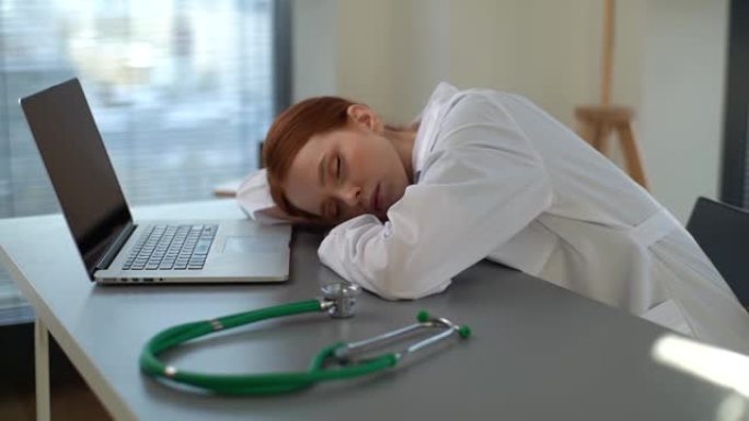 穿着白大褂的疲惫劳累的年轻女医生在工作场所用笔记本电脑和听诊器睡在办公桌前的跟踪镜头。