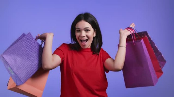 在紫罗兰工作室背景下购物后，兴奋的女人带着彩色纸袋。季节性销售、购买、花钱买礼物的概念