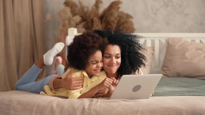 非裔美国妇女和女孩在笔记本电脑键盘上打字，浏览内容和交流。母亲和女儿躺在卧室的床上，室内明亮。慢动作