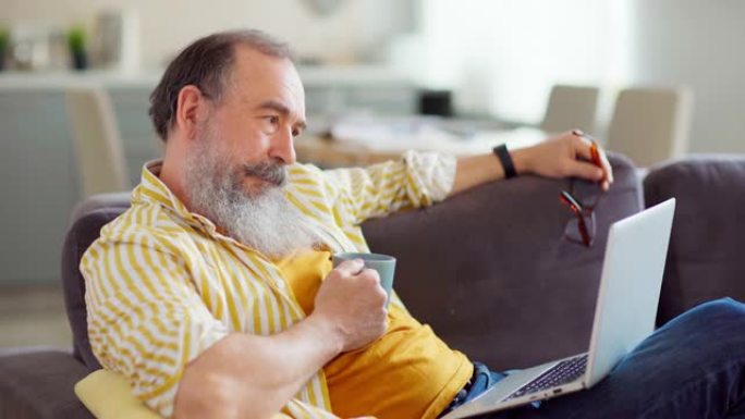 倾斜有思想的高级男人看着笔记本电脑屏幕，叹气，看着相机，继续思考项目坐在家里的沙发上，手里拿着一杯茶