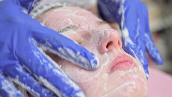 美容师医生正在美容诊所将面部清洁泡沫应用于女性面部按摩皮肤。美容师在美容院制作护肤程序，年轻女子肖像