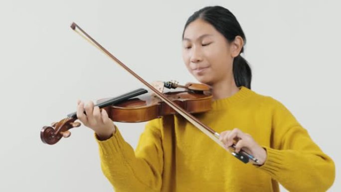 笑着的亚洲女学生穿着黄色毛衣学习拉小提琴与灰色背景，生活理念。