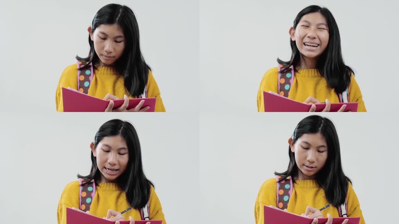 亚洲背包女学生穿着黄色毛衣写在粉红色的书上，在灰色的背景下，生活方式的概念。