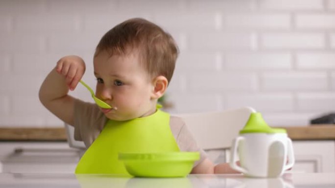 可爱的小男孩坐在桌子旁，用勺子吃燕麦片。宝宝在家心甘情愿地吃东西。概念快乐童年