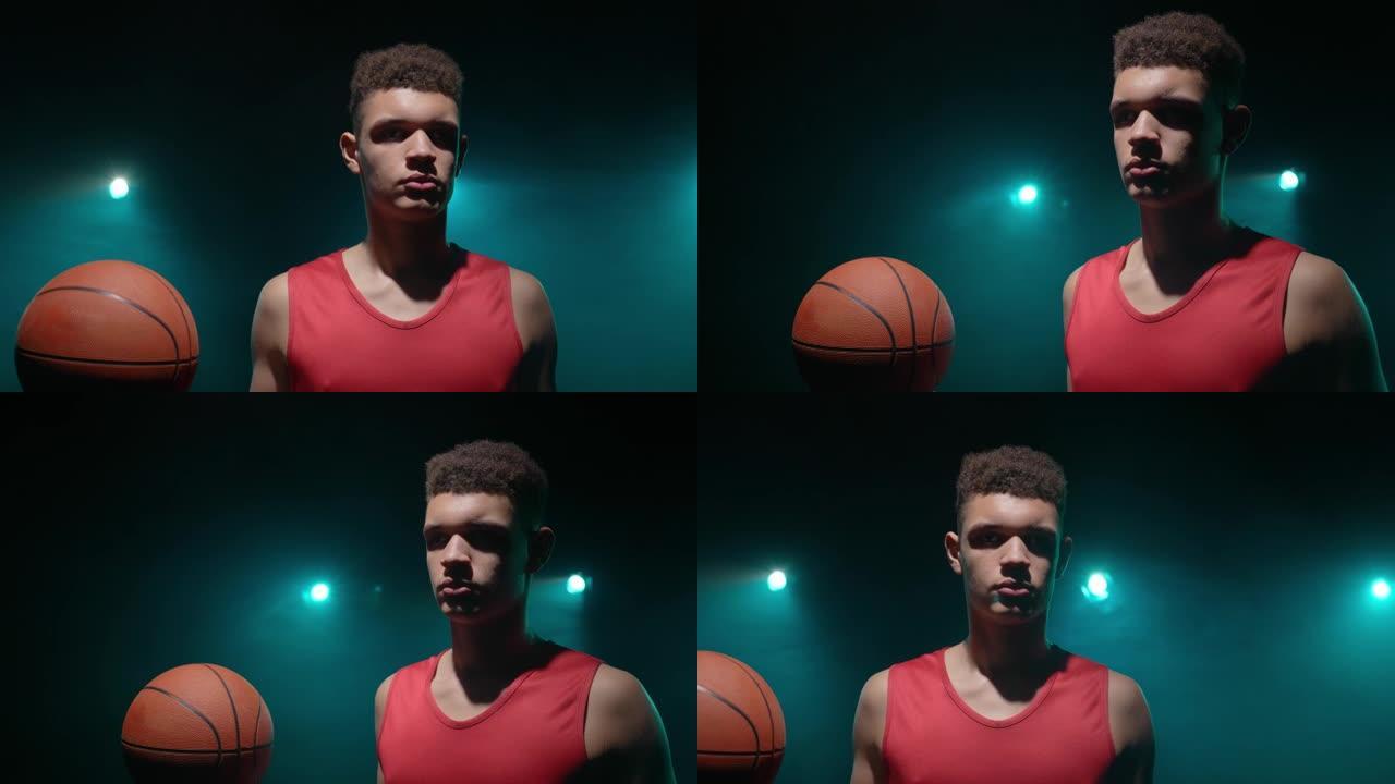 手握球的年轻篮球运动员的肖像。穿着红色运动服的运动员在黑暗的演播室背景上摆姿势，烟熏蓝灯。特写。慢动