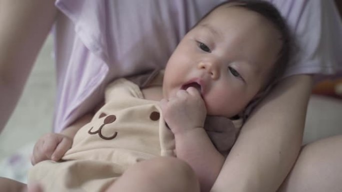 天真的亚洲婴儿婴儿坐在父母母亲身上的肖像。婴儿吮吸手和手指。3个月大的婴儿面部表情。