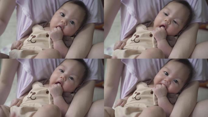 天真的亚洲婴儿婴儿坐在父母母亲身上的肖像。婴儿吮吸手和手指。3个月大的婴儿面部表情。