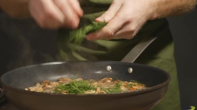 男主厨在热煎锅里将绿莳萝添加到炸蘑菇中。厨师用手摘下莳萝叶，然后将它们和蘑菇一起扔进锅里。