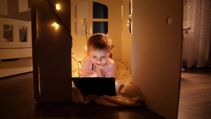 用平板电脑躺在帐篷或纸板玩具屋的地板上的微笑男孩的照片。儿童教育和夜间学习的概念