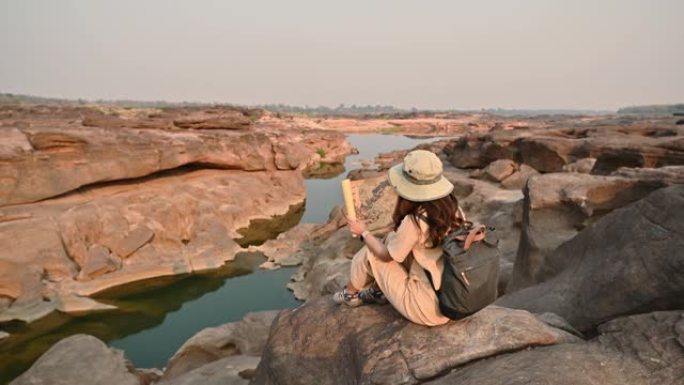 年轻的亚洲女探险家坐在泰国大峡谷岩石悬崖上看纸质地图